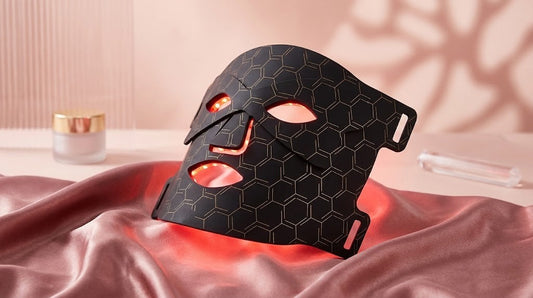 Máscara facial LED de silicona negra con patrón de panal y luces rojas encendidas sobre satén rosa